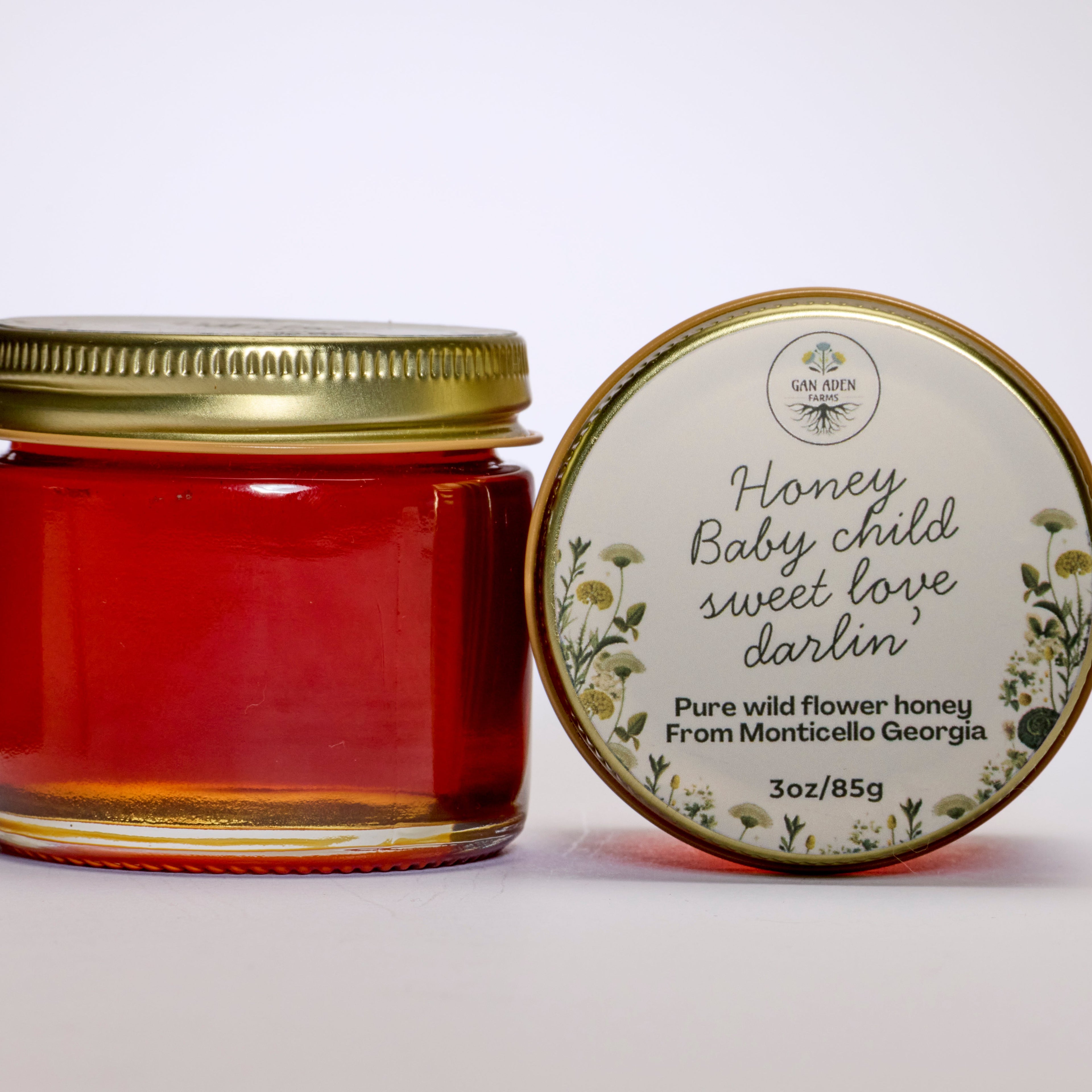 A glass jar of red wildflower honey with a gold metal lid, shown next to its round cream-colored label lid on a clean white background.