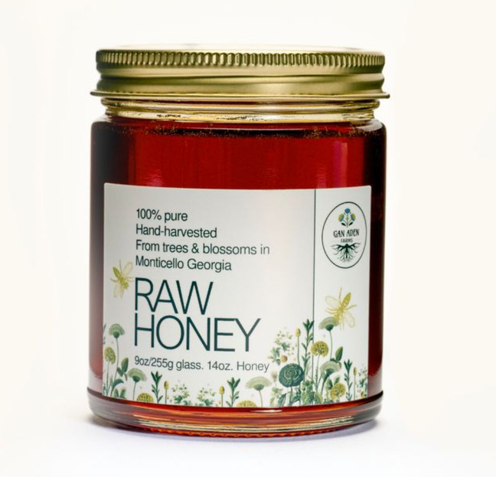 A glass jar of raw honey with a gold metal lid and a white label featuring green text and floral illustrations, set against a plain white background.